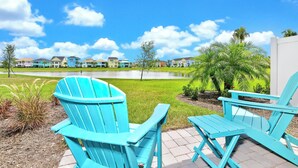 Un espace patio agrémenté de deux fauteuils Adirondack turquoise et d’une petite table, d’où l’on a vue sur un étang et une rangée de maisons aux couleurs vives.