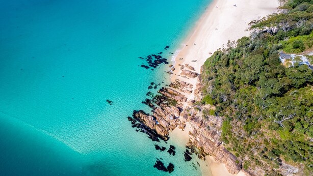 Vista aérea de una playa con aguas turquesas cristalinas, formaciones rocosas y abundante vegetación a la derecha.