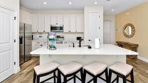 A kitchen with white cabinets, a stainless steel refrigerator, a microwave, and a large island with bar stools.