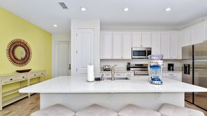 Une cuisine moderne avec des meubles blancs, un grand îlot central doté de tabourets, un four à micro-ondes, un réfrigérateur et un mixeur.