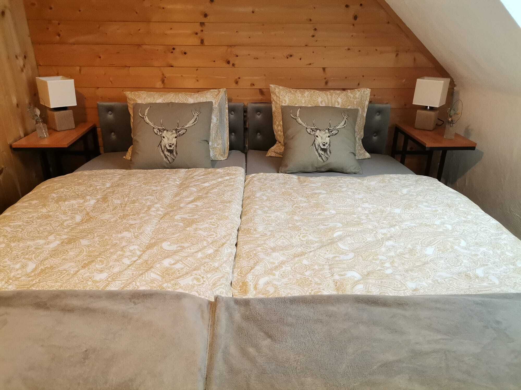 Un dormitorio con techo inclinado de madera, una cama con ropa de cama estampada, dos almohadones decorativos con diseños de ciervo y lámparas de noche sobre mesitas de madera.