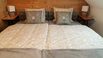 Un dormitorio con techo inclinado de madera, una cama con ropa de cama estampada, dos almohadones decorativos con diseños de ciervo y lámparas de noche sobre mesitas de madera.