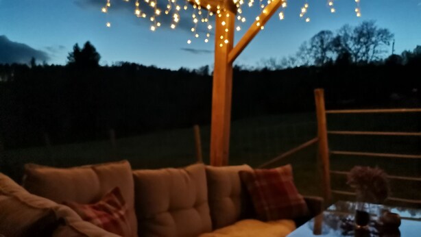 A cozy outdoor seating area with a wooden pergola and string lights, a cushioned sofa, and a glass table with a vase.