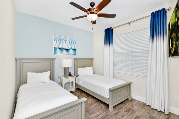A hotel room with two beds, a ceiling fan, a painting of sailboats, and a window with curtains.