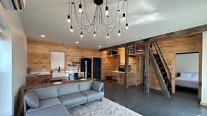 A modern interior with a sectional sofa, wooden walls, a kitchen area, and a staircase. - VRFLX Seneca View Bungalow | Hot Tub Retreat (Burdett)
