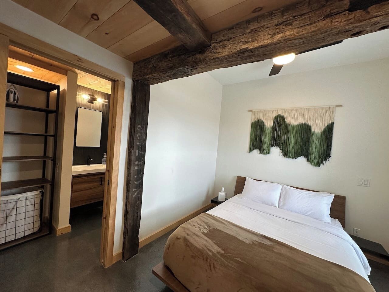 A bedroom with a wooden headboard, a bed with white linens, a nightstand, a wall-mounted artwork, and a bathroom visible through an open door.