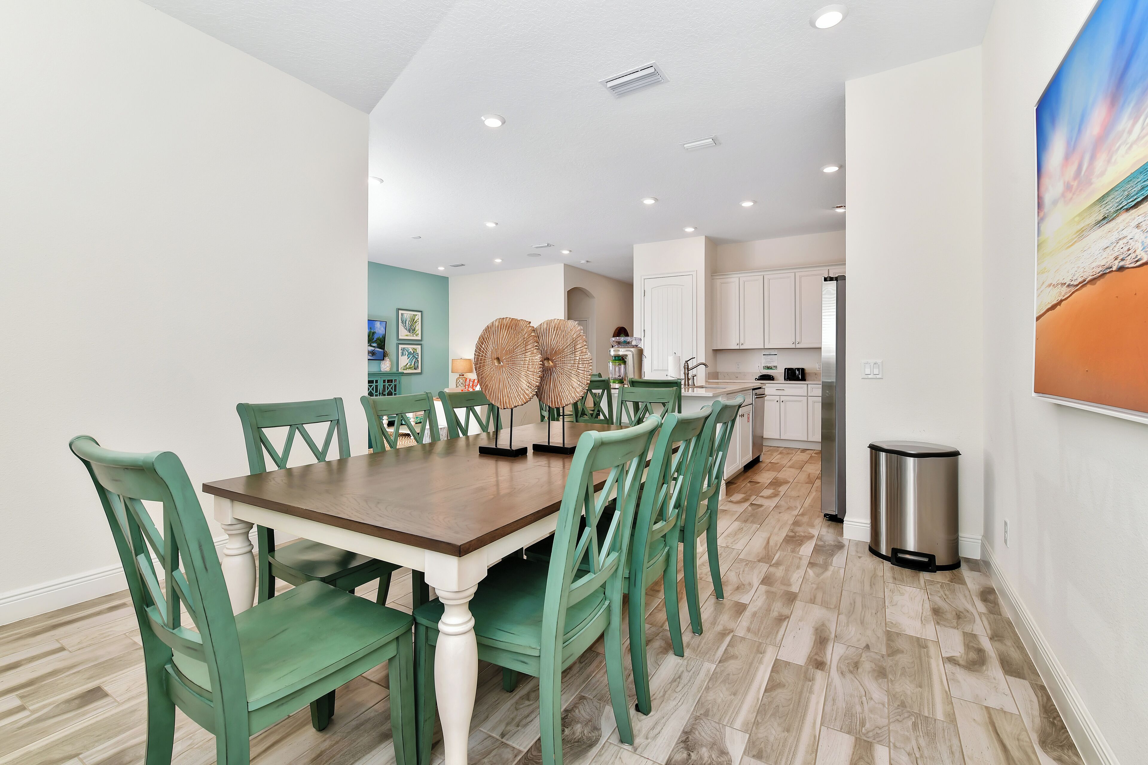 A dining area with a wooden table and green chairs, a kitchen in the background, and a trash can.