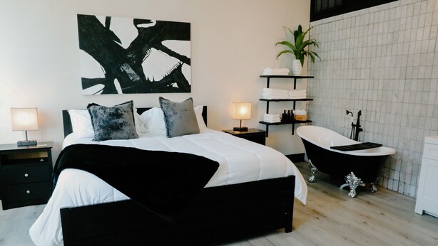 A modern bedroom with a black and white bed, bedside tables with lamps, a black bathtub, and a large abstract painting on the wall.