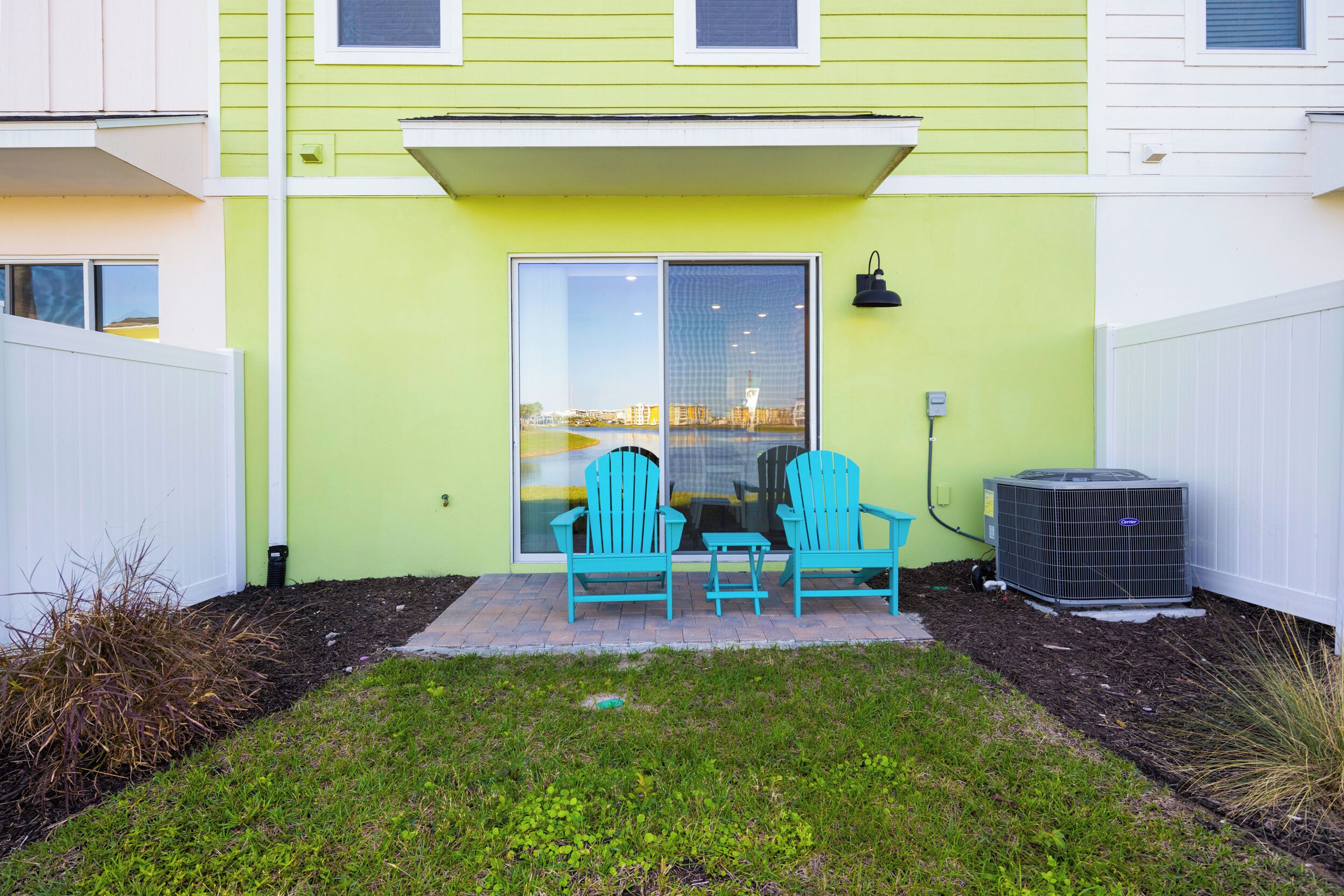 Une terrasse avec deux chaises turquoise, une petite table et un grand climatiseur installé contre le mur d’une maison peinte en vert.
