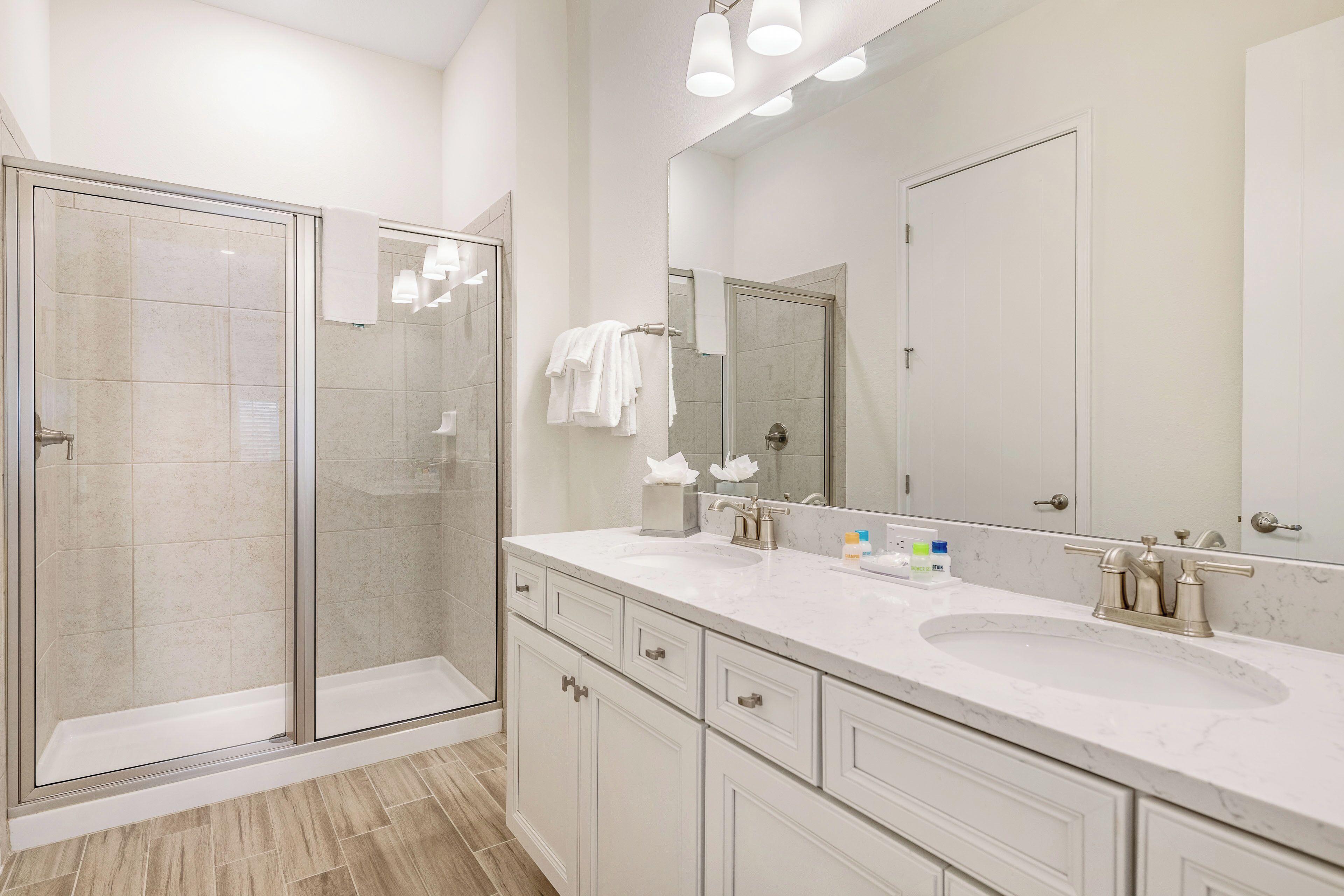 Un baño moderno con mampara de vidrio, un espejo grande y un mueble de tocador blanco con lavabo y varios cajones.