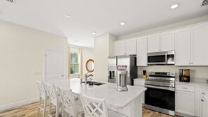 Uma cozinha moderna com armários brancos, uma ilha central com banquetas, eletrodomésticos de aço inoxidável e uma janela com persianas.