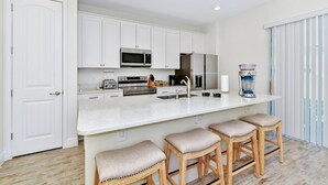 A modern kitchen with white cabinetry, stainless steel appliances, a large island with stools, and a view of the outdoors through a sliding door.