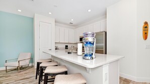 Une cuisine moderne avec des meubles blancs, un îlot central intégré doté de tabourets, un mixeur et une décoration murale représentant une planche de surf.