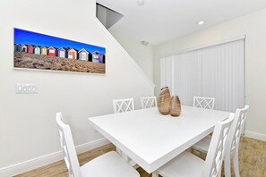 Un comedor con una mesa y sillas blancas, un cuadro con cabañas de playa y una puerta blanca.
