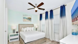 Un dormitorio con cama, mesita de noche, lámpara, ventilador de techo y ventana con cortinas.