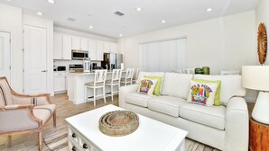 Un salon moderne avec un canapé blanc, une table basse et un coin repas comprenant des chaises blanches, et une cuisine en arrière-plan.