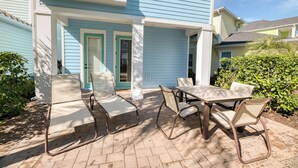 A patio with a table and chairs, a covered area with a door, and a blue house in the background.