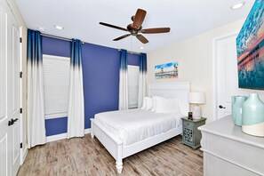 Une chambre à coucher avec un lit blanc, des rideaux bleus, un ventilateur de plafond et un tableau accroché au mur.
