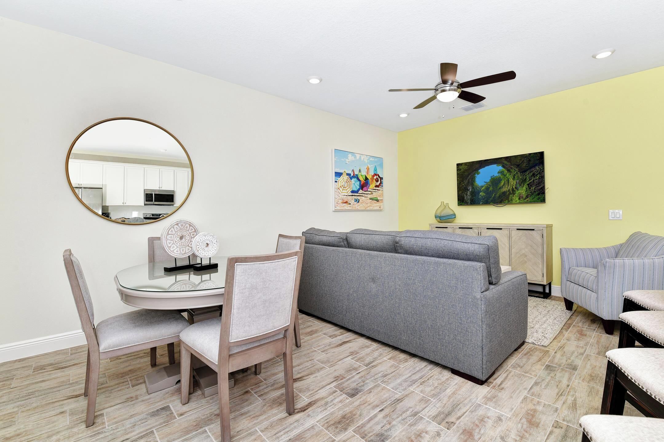A living room with a gray sofa, a dining table with chairs, a round mirror, and a ceiling fan.