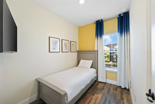 A single bed with a headboard, a window with blue curtains, and three framed pictures on the wall.