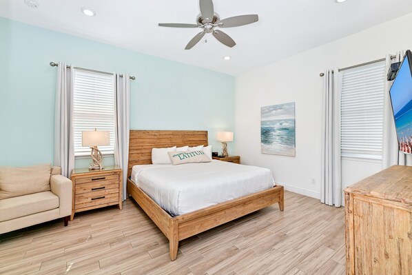 A bedroom with a wooden bed, two bedside tables with lamps, a ceiling fan, and a painting on the wall.