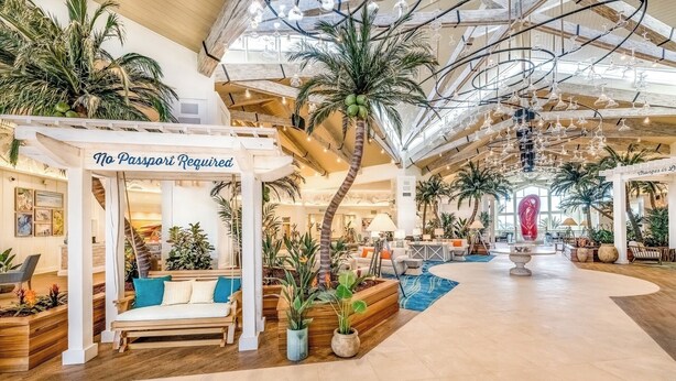 Un vaste atrium intérieur doté d’un haut plafond, avec un espace de détente central agrémenté d’un panneau indiquant « Pas de passeport requis », le tout entouré de plantes en pot et de palmiers.