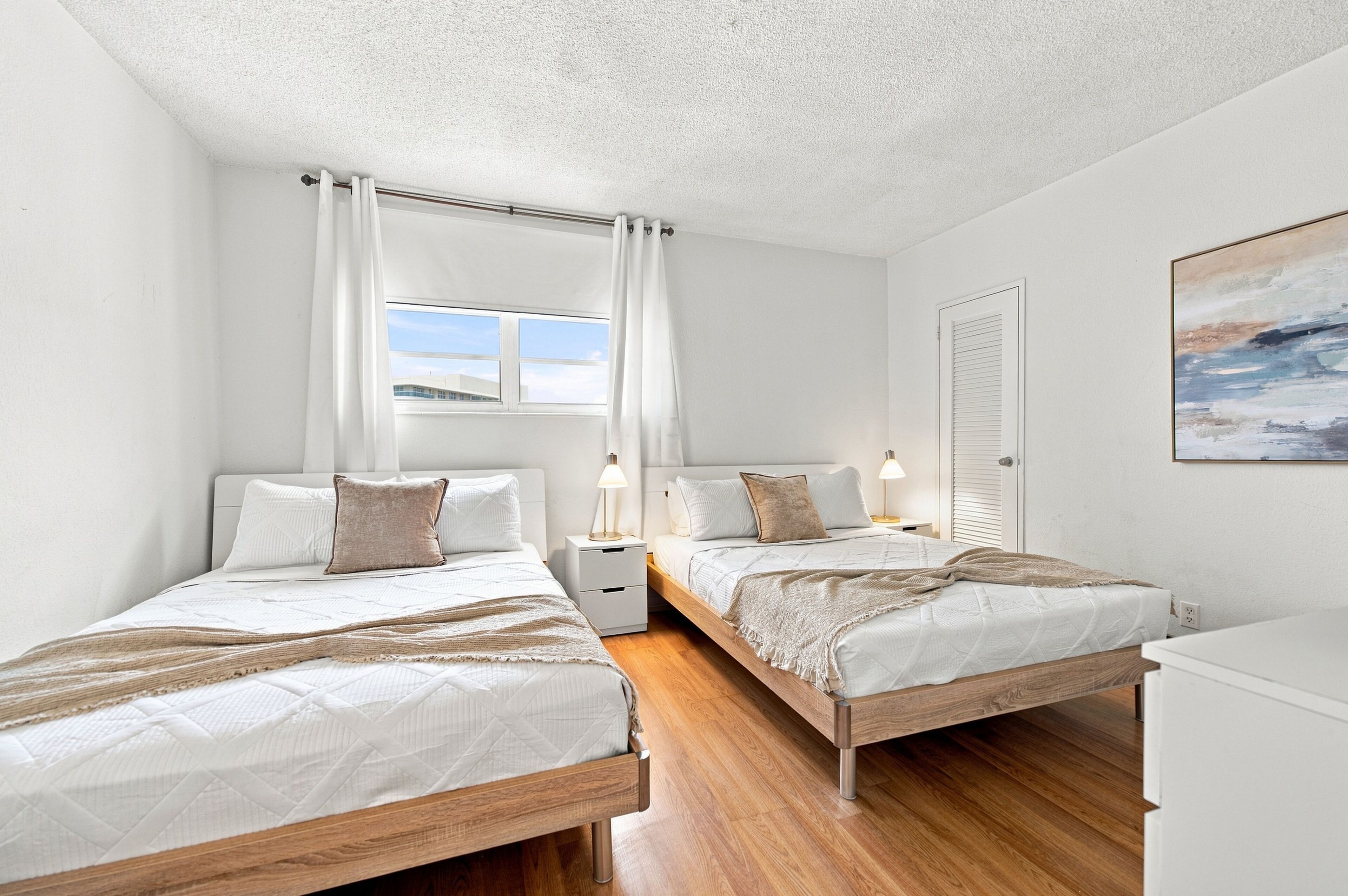 A bedroom with two beds, a window with curtains, bedside lamps, and a painting on the wall.