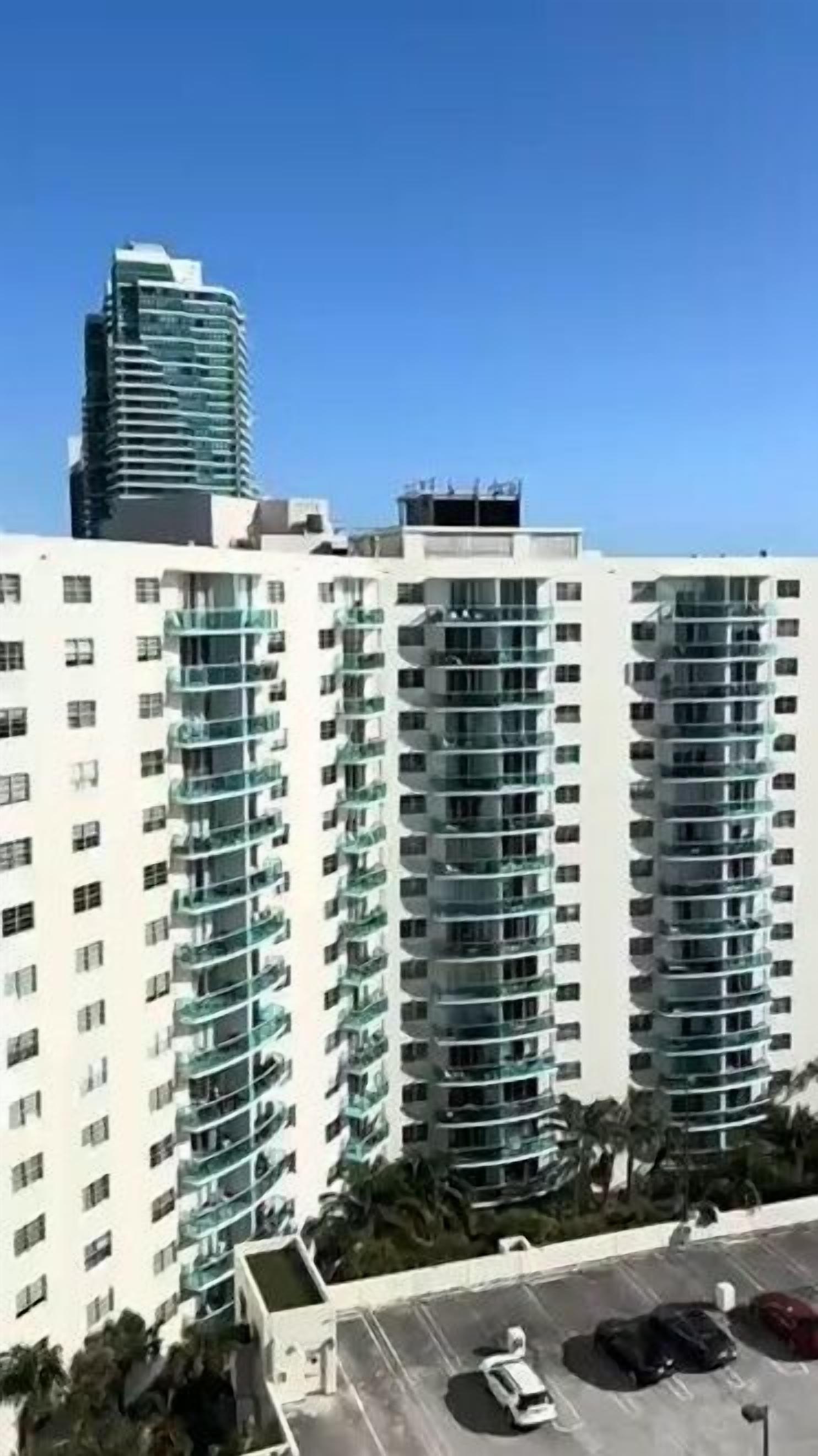 A high-rise residential building with multiple balconies, a parking area in the foreground, and a taller building in the background.
