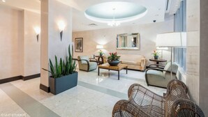 Un hall d’entrée spacieux avec un plafond haut, un espace de détente central avec un canapé et des fauteuils, une grande plante en pot et un miroir fixé au mur.