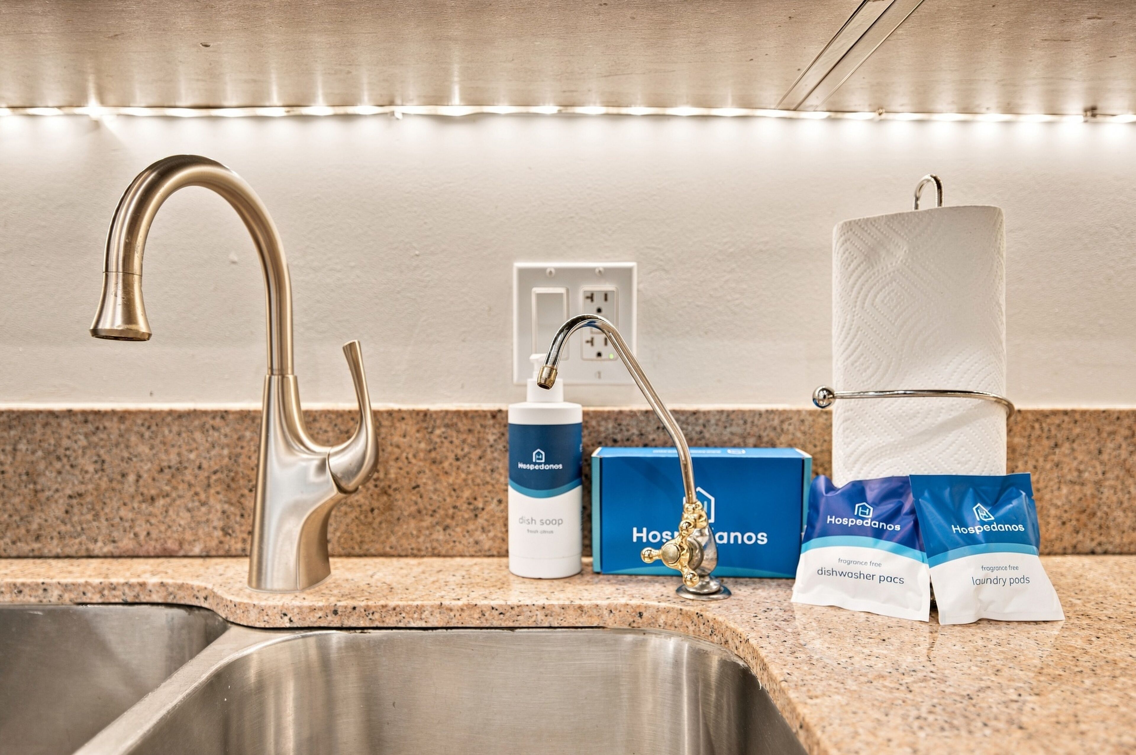 A kitchen sink with a modern faucet, a dish soap bottle, a paper towel dispenser, and two packets of laundry pods.