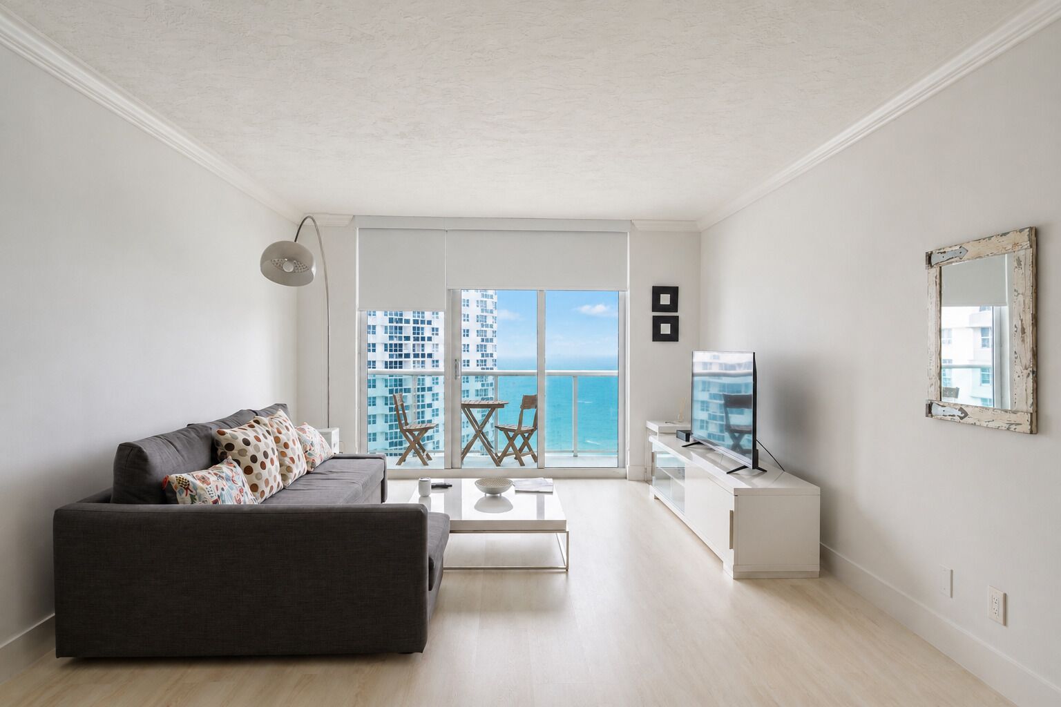 Ein modernes Wohnzimmer mit einem grauen Sofa, einem Couchtisch und einer TV-Konsole. Ein Balkon bietet Blick auf das Meer und die Gebäude.