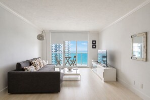 A modern living room with a grey sofa, a coffee table, and a TV stand. There is a balcony with a view of the sea and buildings. - The Tides Building Private Apartments (Hollywood)