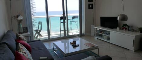 A modern living room with a sofa, coffee table, and TV stand. A balcony with a view of the ocean is visible through large windows.