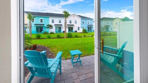 Une vue depuis un balcon, avec des chaises turquoise et une petite table, donnant sur une rangée de maisons colorées, un gazon bien entretenu et des palmiers.