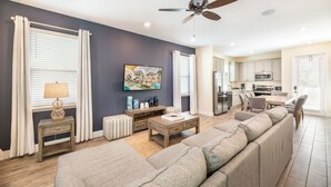 A modern living room with a sectional sofa, a coffee table, and a TV. There is a dining area with a table and chairs, and a kitchen in the background. - Cozy Cottage near Disney | Private Pool + Resort & Waterpark Access - 8036ST (Kissimmee)
