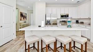 Una cocina moderna con armarios blancos, una isla central con taburetes, un refrigerador, un microondas y una licuadora.