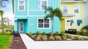Un immeuble résidentiel de deux étages, avec une façade turquoise et jaune, un palmier et un banc dans le jardin.
