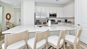 A modern kitchen with white cabinetry, a large island with bar stools, stainless steel appliances, and a wooden dining table.