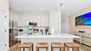 Une cuisine moderne avec des meubles blancs, un grand îlot central doté de tabourets, des appareils en acier inoxydable et un tableau représentant une scène de plage accroché au mur.