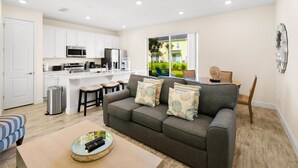 Ein modernes Wohnzimmer mit grauem Sofa, einem Couchtisch und einem Fernseher. Es gibt eine Küchenzeile mit weißen Schränken und einer Mikrowelle. Ein Deckenventilator ist sichtbar, und das Zimmer hat große Fenster.