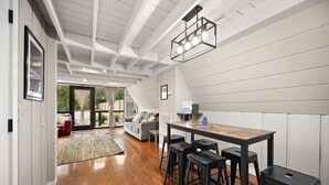 Una cocina moderna con una mesa de madera y taburetes de bar, un ventanal con vistas al exterior, y un techo con vigas a la vista e iluminación empotrada.