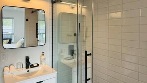A modern bathroom with a glass shower enclosure, a white sink with a mirror, and a towel rack. - Brand New Studio with Pool Access (Oakland Park)