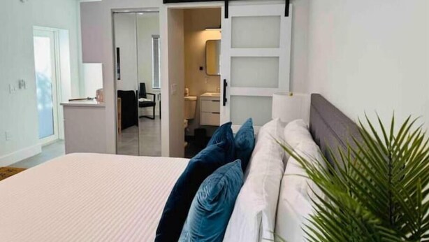 A bedroom with a bed, a sliding door leading to a bathroom, and a plant in the corner. - Brand New Studio with Pool Access (Oakland Park)