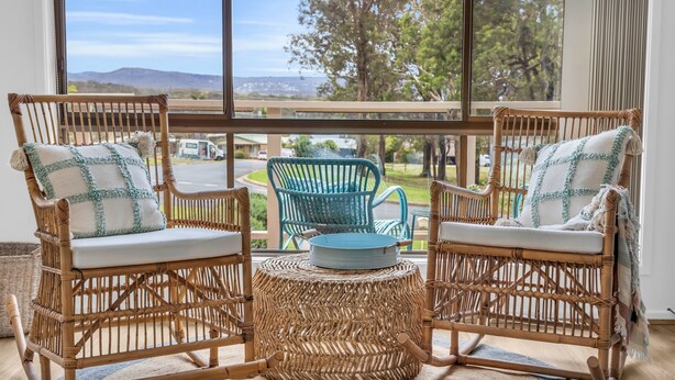 Two wicker chairs with white cushions and a woven stool in a room with large windows overlooking a road and greenery. - Seaglass at Pambula Beach (Pambula Beach)