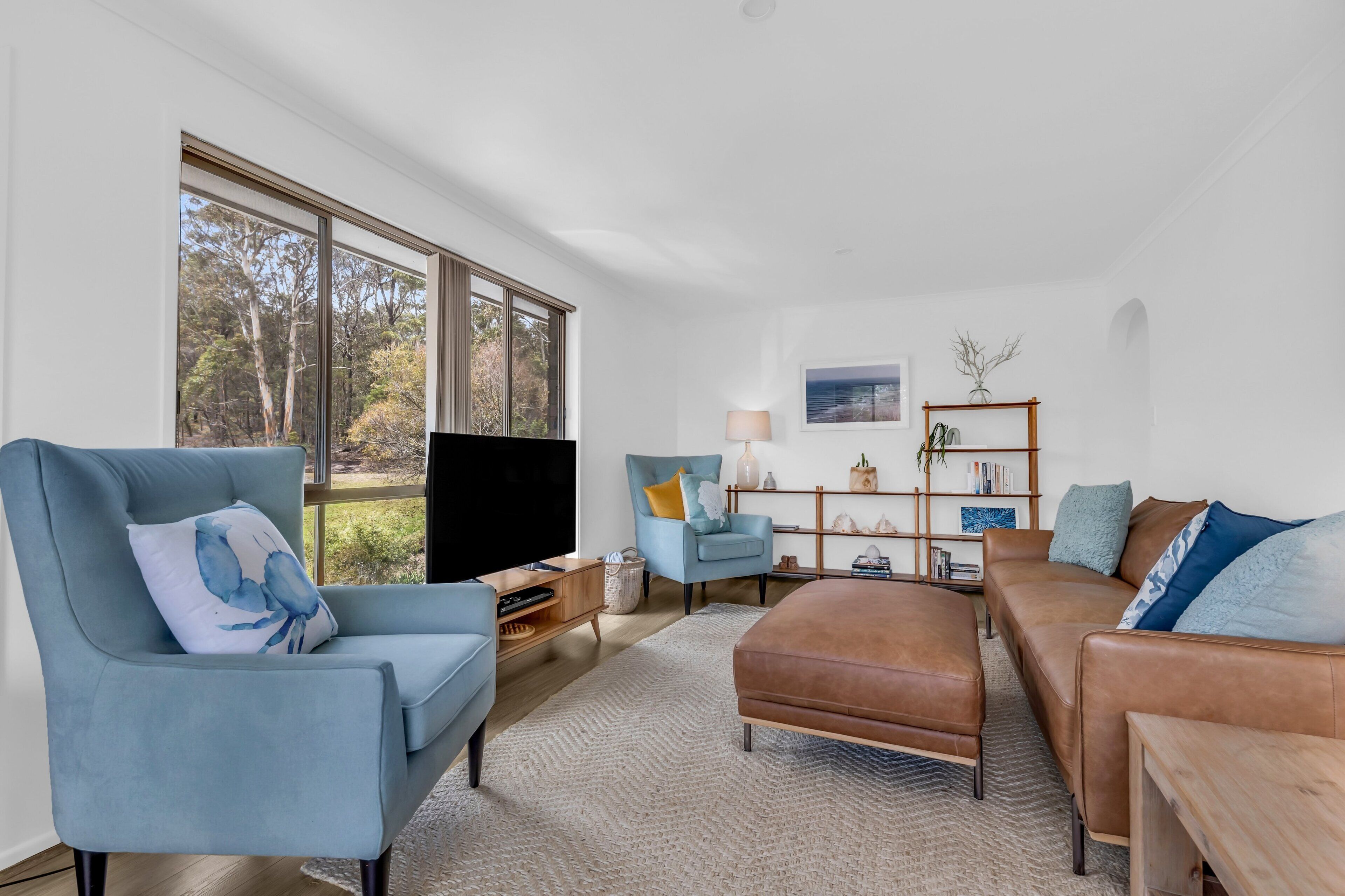 A modern living room with a large window, a blue armchair, a brown sofa, and a wooden side table.