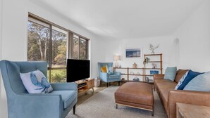 A modern living room with a large window, a blue armchair, a brown sofa, and a wooden side table. - Seaglass at Pambula Beach (Pambula Beach)
