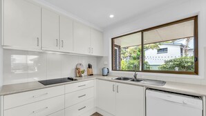 A modern kitchen with white cabinets, a built-in oven, a dishwasher, and a window overlooking a residential area. - Seaglass at Pambula Beach (Pambula Beach)