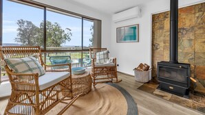 A cozy living room with wicker furniture, a stone fireplace, and a large window overlooking a scenic view. - Seaglass at Pambula Beach (Pambula Beach)