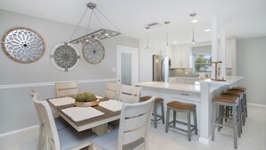 A modern kitchen with white cabinetry, a central island with stools, and decorative wall art. - Vandy Villas — Walk to Vanderbilt Beach, Pool & Spa (Naples)