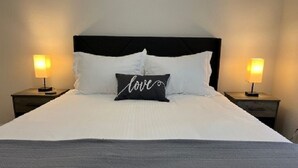 Ein ordentlich bezogenes Bett mit weißen Leinentüchern und einem schwarzen Kopfkissen mit dem Wort "love". Auf beiden Seiten des Bettes stehen je eine Nachttischlampe.